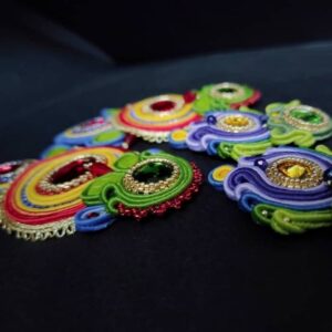 Sullo sfondo nero si vede dettagli di due paia di orecchini colorati estivi fatti a mano con la tecnica di soutache, cristalli verdi, rossi e gialli, perline dorate.