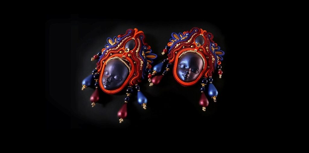 Orecchini artigianali in soutache con volti scultorei in resina blu, incorniciati da fili intrecciati nei toni del rosso, arancione e blu. Decorati con perline dorate, cristalli e pendenti satinati bordeaux e blu notte, questi gioielli unici evocano un fascino mistico e teatrale.